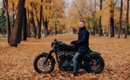 Biker with thick beard, wears protective gloves, black coat and jeans, poses on motobike in park
