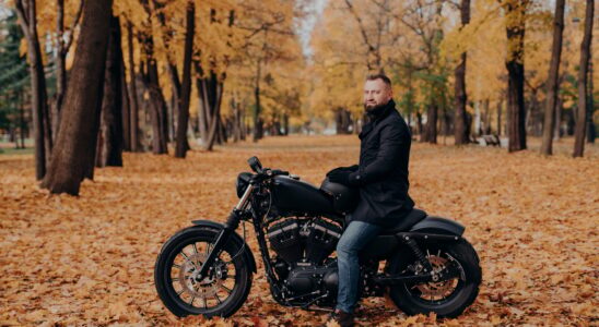 Biker with thick beard, wears protective gloves, black coat and jeans, poses on motobike in park